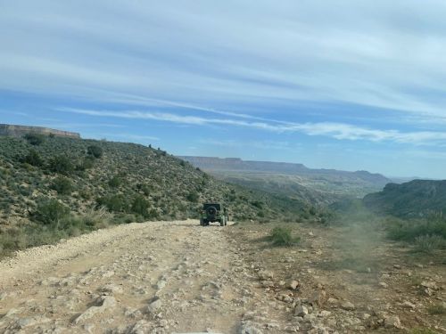 A rugged dirt road winds through a rocky landscape, with a vehicle driving away into the distance under a cloudy sky.