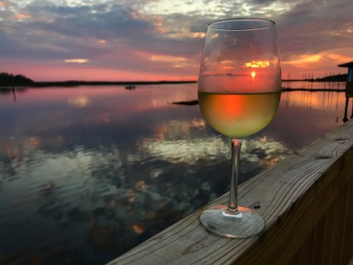 A glass of white wine sits on a wooden railing, reflecting a colorful sunset over a calm water surface.