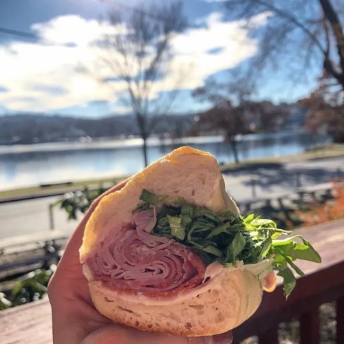 A hand holding a sandwich with layers of ham and lettuce, set against a scenic lakeside background.