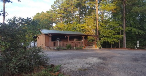 A cozy cabin surrounded by trees, featuring a porch and gravel driveway, set in a serene, wooded area.