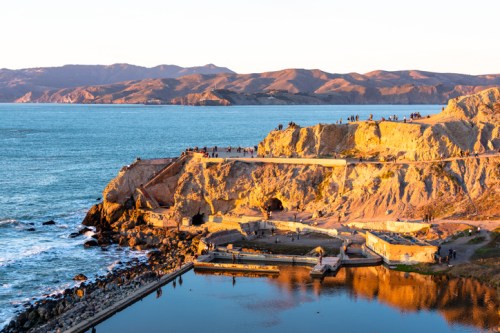 A scenic view of a rocky coastline at sunset, with people on the cliffs and mountains in the background.