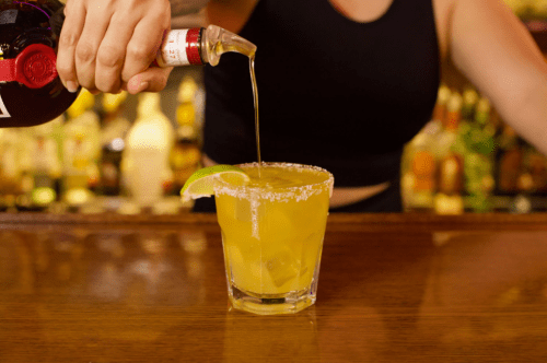 A bartender pours a drink into a salted glass with a lime wedge, set against a blurred bar background.