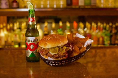 A burger with toppings and fries in a basket, alongside a bottle of Dos Equis beer, set against a blurred bar background.