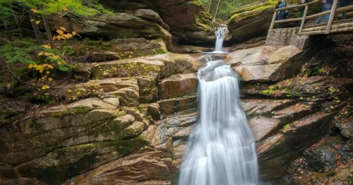 A serene waterfall cascades over rocky cliffs, surrounded by lush greenery and autumn leaves.