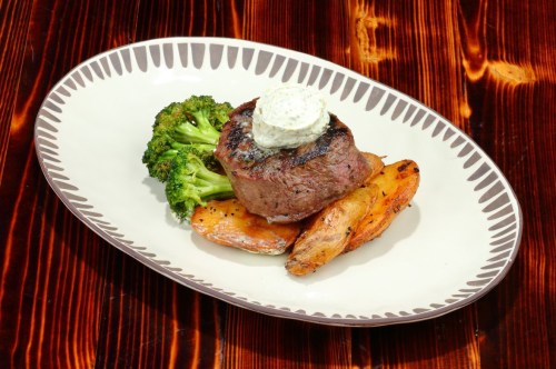 A plated meal featuring a grilled steak topped with herb butter, served with broccoli and roasted potatoes.
