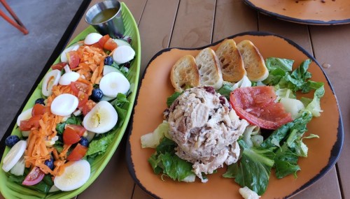A colorful salad with eggs, tomatoes, and blueberries next to a plate of chicken salad on a bed of lettuce with toast.