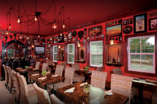 Cozy restaurant interior with red walls, wooden tables, woven chairs, and an array of framed artwork and photographs.