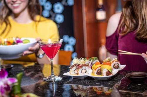 A vibrant sushi platter and a pink cocktail on a table, with two women enjoying their meal in a stylish setting.