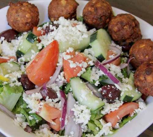 A fresh salad with tomatoes, cucumbers, onions, olives, feta cheese, and falafel balls on the side.