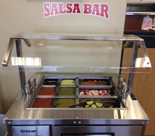 A salsa bar with various salsas and toppings displayed in metal containers under a clear cover.