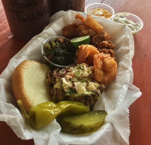 A plate featuring fried shrimp, coleslaw, pickles, a bun, and sides of greens and sauces in small containers.