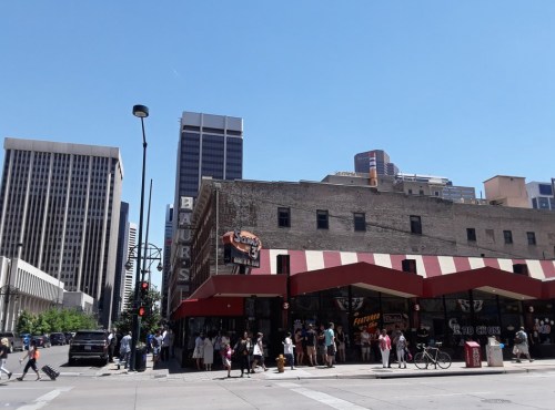 A bustling city street with a historic building, people walking, and tall skyscrapers in the background under a clear blue sky.