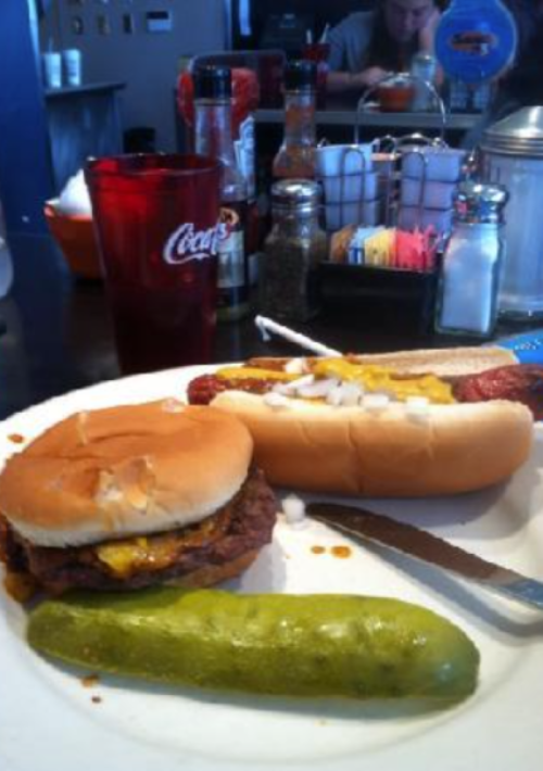 A plate with a cheeseburger, hot dog topped with onions, and a pickle, set in a diner with condiments in the background.