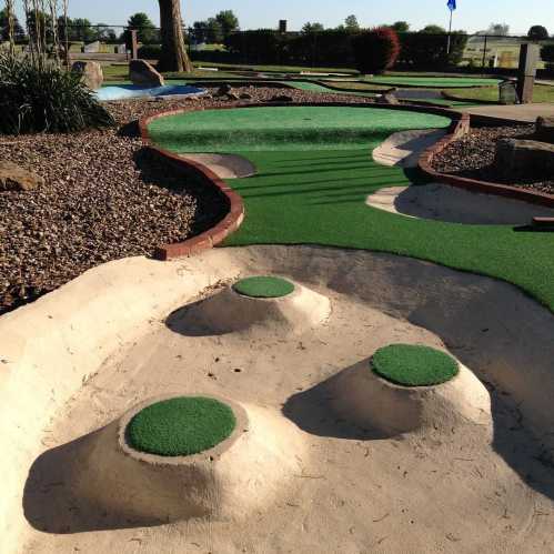 A mini-golf course featuring a winding path with three small mounds and green turf, surrounded by rocks and grass.