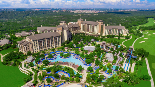 Aerial view of a luxury resort with pools, gardens, and golf course, surrounded by lush green hills and blue skies.