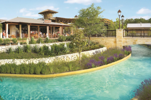 A scenic view of a restaurant by a winding blue waterway, surrounded by greenery and purple flowers under a clear sky.