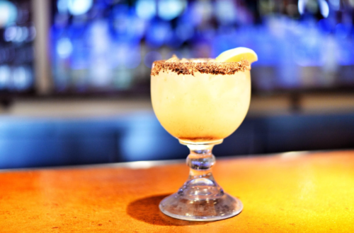 A margarita in a glass with a salted rim, garnished with a lime wedge, on a bar counter with a blurred background.