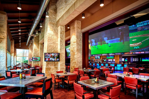 A spacious restaurant with stone walls, red chairs, and large screens showing sports events.