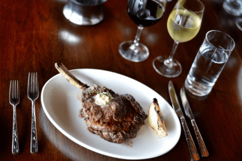 A plated steak with a pat of butter, accompanied by wine glasses and a glass of water on a wooden table.