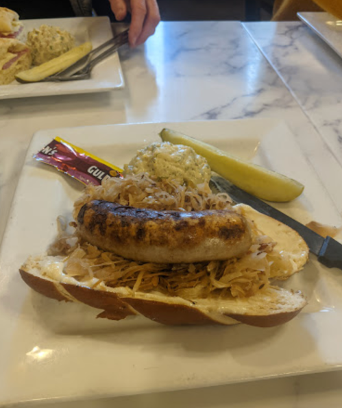 A grilled sausage on a bun topped with sauerkraut, served with a pickle and a packet of mustard on a white plate.
