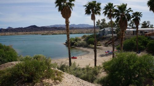 A serene river scene with palm trees, a sandy beach, and kayaks near a calm waterway surrounded by mountains.