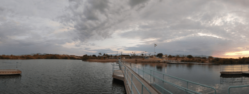 A panoramic view of a calm lake at sunset, with a dock extending into the water and clouds in the sky.