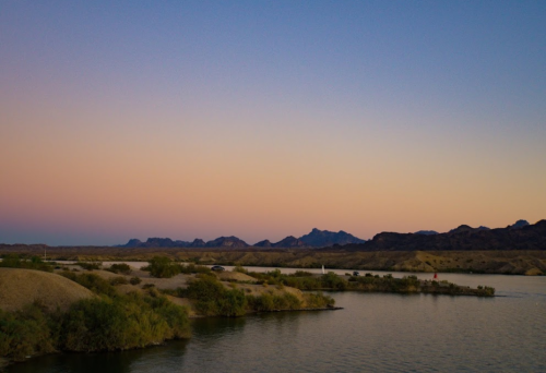 A serene landscape at dusk, featuring a river, sandy banks, and distant mountains under a colorful sky.