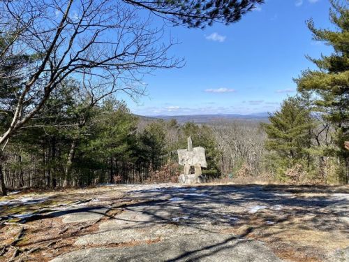 A scenic view from a rocky overlook, featuring trees and distant mountains under a clear blue sky.