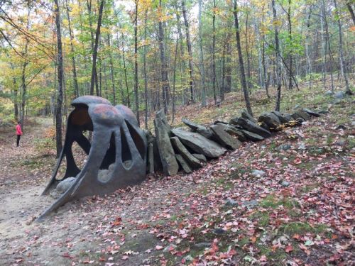A large, abstract sculpture resembling a creature, surrounded by rocks, in a forest with autumn foliage.