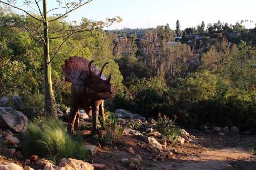 A triceratops statue stands among greenery and rocks in a natural setting, with trees and hills in the background.