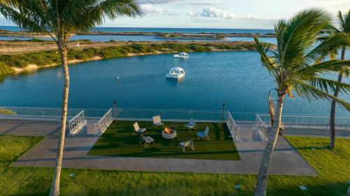 A serene waterfront scene with palm trees, a fire pit, and boats on calm water under a clear sky.