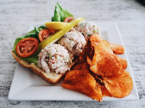 A sandwich with chicken salad, lettuce, tomatoes, and pickles, served with a side of crispy potato chips.