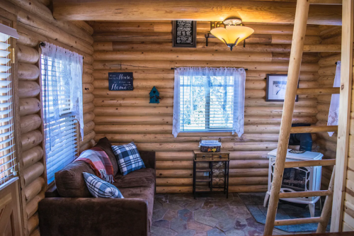 Cozy log cabin interior with a brown couch, decorative pillows, and a small desk by the window. Warm wooden walls.