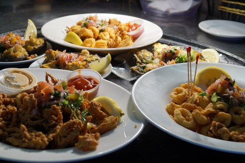 A variety of fried seafood dishes on white plates, garnished with lemon and fresh toppings, served with dipping sauces.