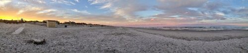 A panoramic view of a beach at sunset, with soft waves and a colorful sky filled with clouds.