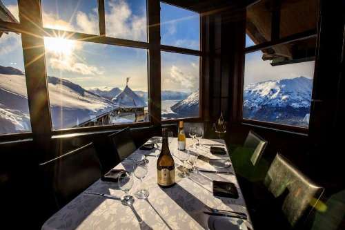 A dining table set with wine and glasses, overlooking a snowy mountain landscape through large windows.