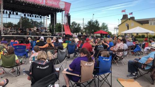 A crowd enjoys a music festival, seated in folding chairs, with a stage and tents in the background.