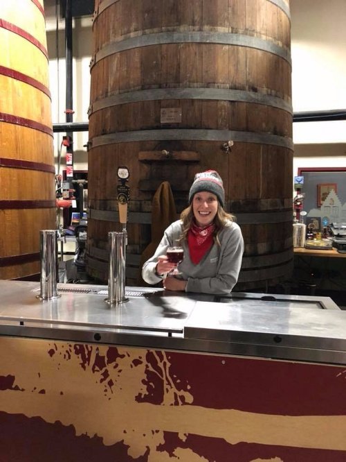 A smiling woman in a gray sweater and beanie holds a drink at a bar with large wooden barrels in the background.