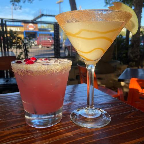 Two colorful cocktails on a wooden table: a pink drink with a sugared rim and a yellow drink with a twist.