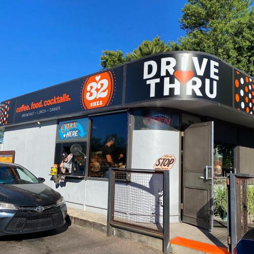 A drive-thru café with a sign reading "32 Shea" offering coffee, food, and cocktails, surrounded by greenery.