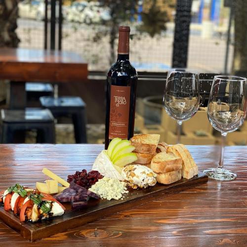 A wooden platter with cheese, fruits, and bread, accompanied by a bottle of wine and two glasses.