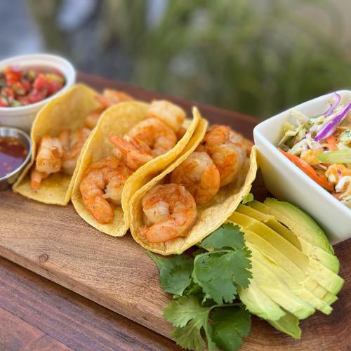 Three shrimp tacos on a wooden platter, accompanied by salsa, avocado slices, and a side of coleslaw.