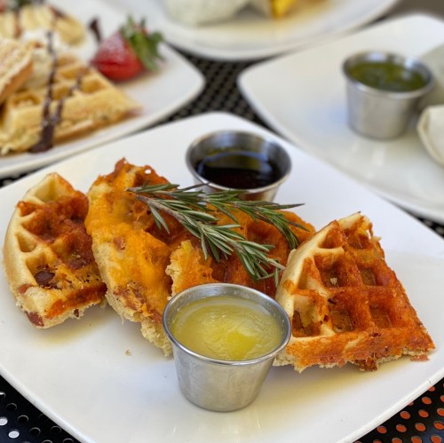 Three savory waffles topped with herbs, served with a small cup of syrup and a citrus sauce on a white plate.