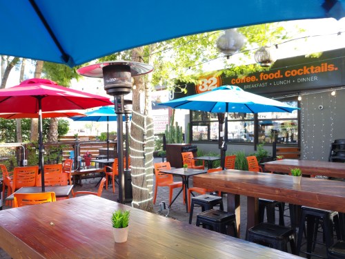 Outdoor dining area with colorful umbrellas, wooden tables, and orange chairs, surrounded by greenery and string lights.