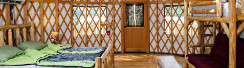 Cozy interior of a yurt with wooden accents, featuring beds, a ladder to a loft, and large windows.
