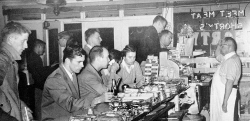 A black-and-white photo of men seated at a diner counter, with a server behind the counter and a bustling atmosphere.