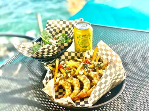 A plate of shrimp tacos with toppings, fries, and a can of soda, alongside a bowl of salad, set against a scenic backdrop.