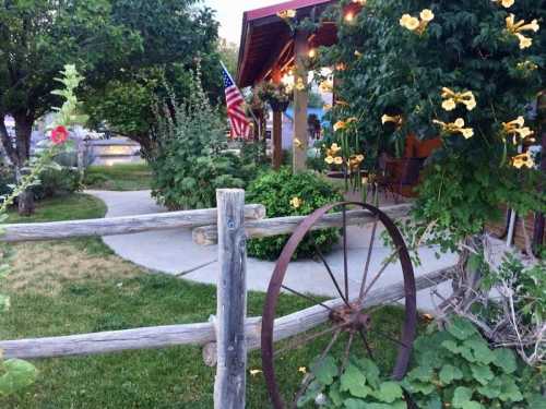 A rustic scene featuring a wooden fence, a vintage wheel, and vibrant flowers near a cozy building with an American flag.