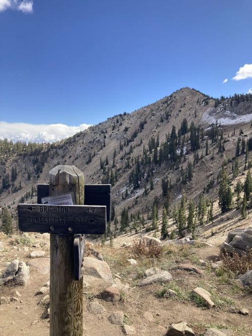 A wooden trail sign points to Brighton and Sunset Pass, with a mountainous landscape and blue sky in the background.