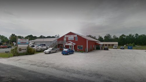 A red building with a sign, surrounded by a gravel parking lot and trees, houses a restaurant and bar.